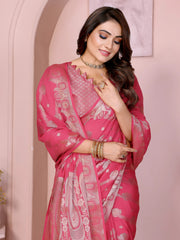Woman wearing a pink Banarasi saree featuring intricate gold zari motifs and a decorative border with tassel details, paired with a matching blouse with short sleeves. She stands gracefully in front of a pastel arched backdrop, styled with traditional jewelry and bangles, with minimal decor including a white pedestal with vases and a potted green plant, showcasing a festive ethnic look.