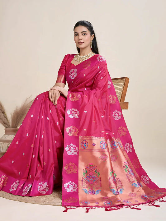 Woman wearing a vibrant pink Banarasi silk saree adorned with intricate gold and multicolor woven motifs and a contrasting gold border, paired with a matching blouse and traditional jewelry, elegantly posing beside a wooden chair in a minimalist studio setting with neutral decor, ceramic vases, and pampas grass.