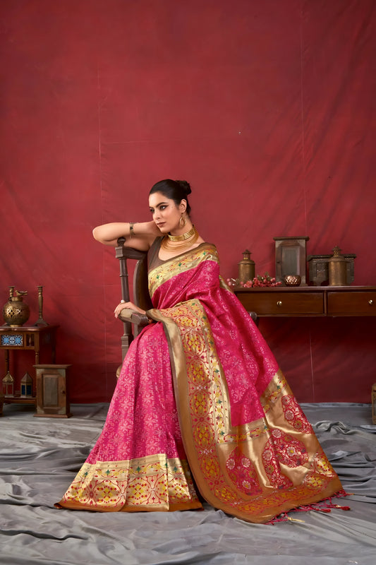 Woman wearing a pink Banarasi silk saree with intricate gold zari border and traditional motifs, accessorized with gold jewelry, standing in a vintage-inspired setting with wooden furniture and a deep red backdrop.
