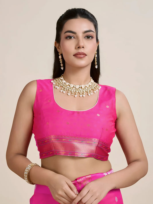 Woman wearing a vibrant pink Banarasi silk saree adorned with intricate gold and multicolor woven motifs and a contrasting gold pallu, paired with a matching blouse and traditional jewelry, elegantly posing beside a wooden chair in a minimalist studio setting with neutral decor, ceramic vases, and pampas grass.