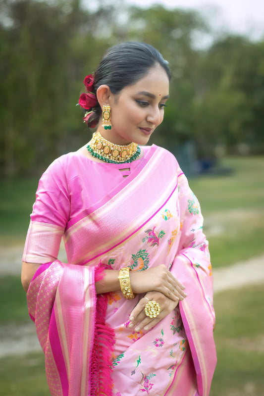  Woman wearing a pastel pink silk saree with floral prints and a decorative border, accessorized with traditional gold jewelry and bangles, standing elegantly on a grassy garden path surrounded by lush greenery and trees.