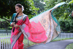 Woman wearing a vibrant pink Madhubani-printed saree with intricate traditional motifs and a contrasting green blouse, accessorized with jewelry and a long braid, standing gracefully on a garden path lined with a white fence and surrounded by lush green foliage.