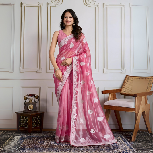 Woman wearing a pastel pink organza saree embellished with delicate white floral embroidery and a matching embroidered border, paired with a sleeveless blouse. She stands elegantly in a classic white paneled room featuring a vintage rotary telephone on a wooden table, a woven chair with a wooden frame, and an ornate patterned rug, creating a sophisticated traditional ambiance.