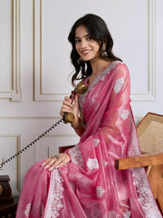 Woman wearing a pastel pink organza saree embellished with delicate white floral embroidery and a matching embroidered border, paired with a sleeveless blouse. She stands elegantly in a classic white paneled room featuring a vintage rotary telephone on a wooden table, a woven chair with a wooden frame, and an ornate patterned rug, creating a sophisticated traditional ambiance.