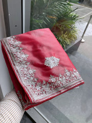 Woman wearing a pastel pink organza saree embellished with delicate white floral embroidery and a matching embroidered border, paired with a sleeveless blouse. She stands elegantly in a classic white paneled room featuring a vintage rotary telephone on a wooden table, a woven chair with a wooden frame, and an ornate patterned rug, creating a sophisticated traditional ambiance.