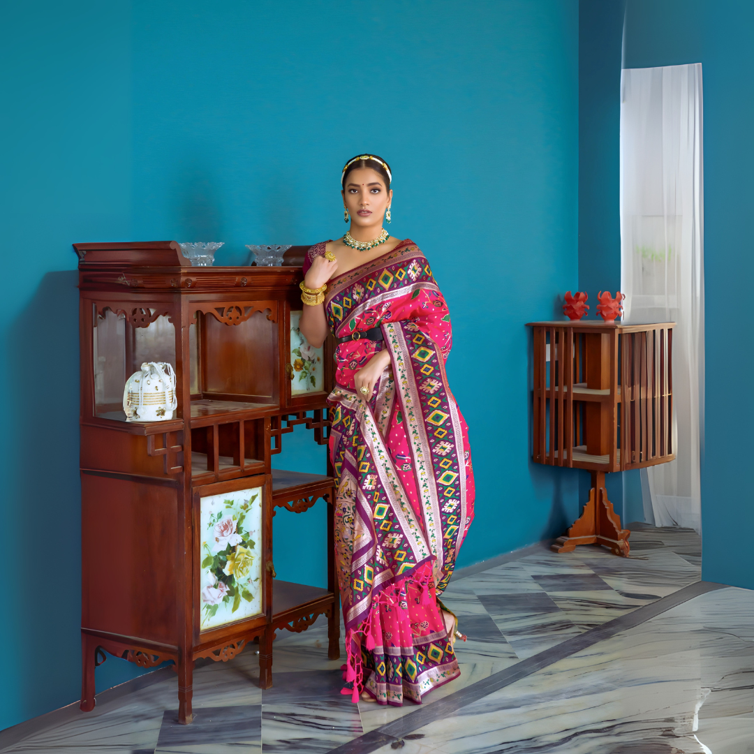 Woman wearing a vibrant pink Patola silk saree with intricate multicolor geometric patterns and a contrasting border, accessorized with traditional gold jewelry, elegantly posing beside vintage wooden furniture in a stylish room with teal blue walls, decorative ceramics, and marble flooring.