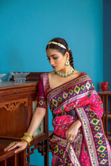 Woman wearing a vibrant pink Patola silk saree with intricate multicolor geometric patterns and a contrasting border, accessorized with traditional gold jewelry, elegantly posing beside vintage wooden furniture in a stylish room with teal blue walls, decorative ceramics, and marble flooring.