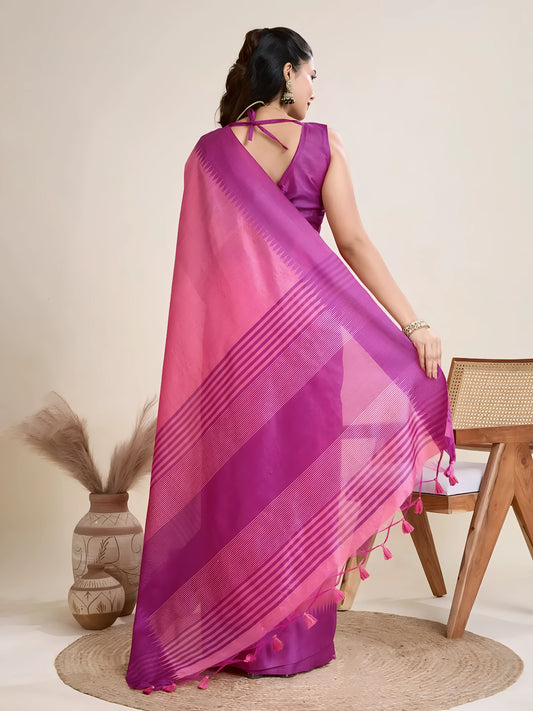 Woman wearing a pink and purple silk saree with a bold striped border and pallu, paired with a matching blouse and traditional jewelry, elegantly posing beside a wooden chair in a minimalist studio setting with neutral decor, ceramic vases, and pampas grass.