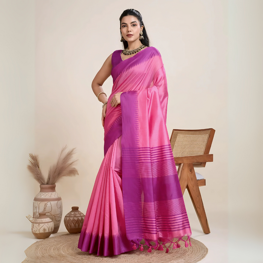 Woman wearing a pink and purple silk saree with a bold striped border and pallu, paired with a matching blouse and traditional jewelry, elegantly posing beside a wooden chair in a minimalist studio setting with neutral decor, ceramic vases, and pampas grass.