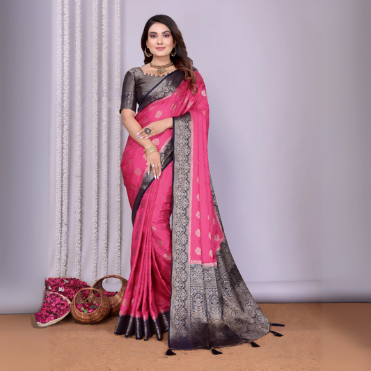 Woman wearing an elegant pink silk saree with a striking black and silver zari border and intricate woven motifs, paired with a black blouse and accessorized with traditional jewelry. She poses gracefully in a modern studio with woven baskets of rose petals and a neutral background, complemented by sheer white curtains for a refined, festive look.
