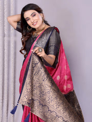 Woman wearing an elegant pink silk saree with a striking black and silver zari border and intricate woven motifs, paired with a black blouse and accessorized with traditional jewelry. She poses gracefully in a modern studio with woven baskets of rose petals and a neutral background, complemented by sheer white curtains for a refined, festive look.