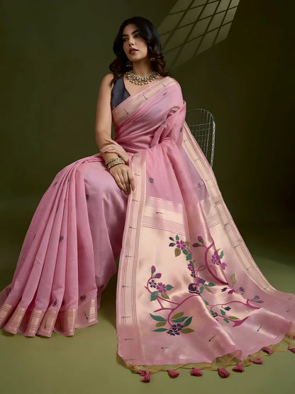 Woman wearing a pastel pink silk saree with subtle woven motifs and a contrasting pallu adorned with floral embroidery, paired with a sleeveless blouse and traditional jewelry, gracefully posing in a studio with olive green walls and patterned window light.Woman wearing a pastel pink silk saree with subtle woven motifs and a contrasting pallu adorned with floral embroidery, paired with a sleeveless blouse and traditional jewelry, gracefully posing in a studio with olive green walls and patterned window.