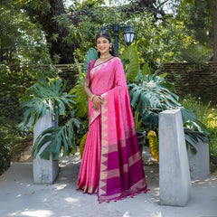 Woman wearing a vibrant pink silk saree with a contrasting magenta and gold zari border, paired with traditional jewelry, standing gracefully outdoors amidst lush green tropical plants and garden decor.