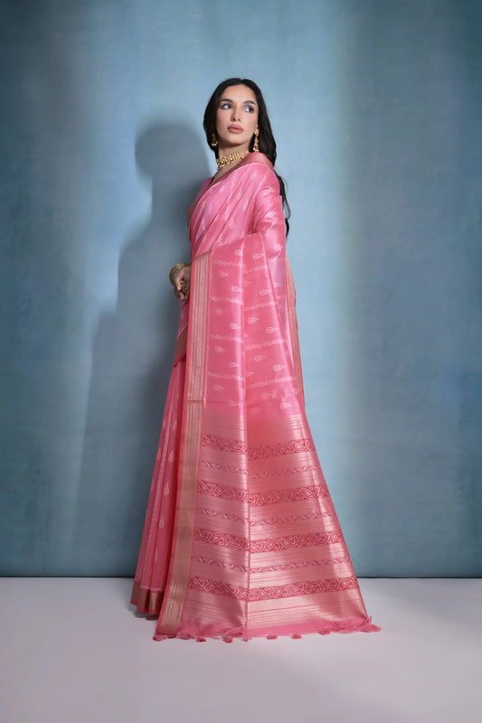 Woman wearing a pink silk saree with intricate woven patterns and gold border, paired with a matching blouse and traditional gold jewelry including a choker necklace and earrings, posing elegantly in a studio setting with a soft blue-grey gradient background.