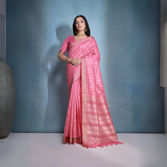 Woman wearing a pink silk saree with intricate woven patterns and gold border, paired with a matching blouse and traditional gold jewelry including a choker necklace and earrings, posing elegantly in a studio setting with a soft blue-grey gradient background.