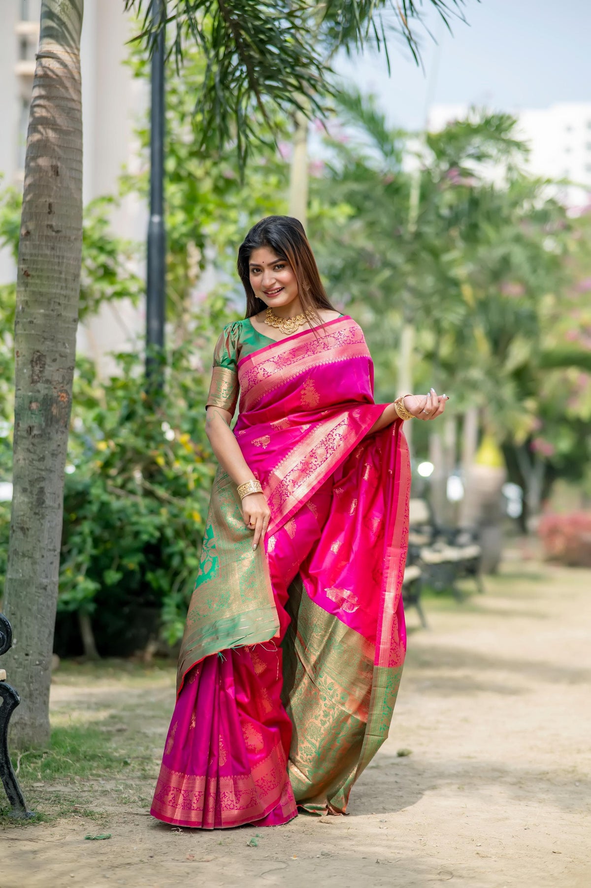 Woman wearing a vibrant pink zari work saree and gold jewelry smiling outdoors in a lush garden setting