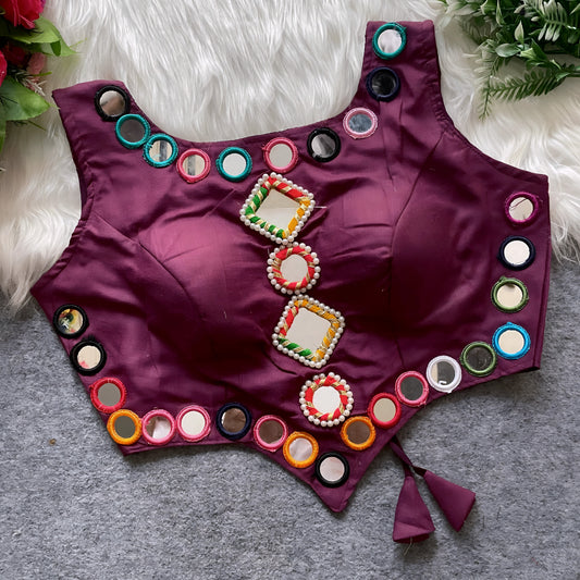 Plum purple ethnic blouse featuring intricate mirror work and multicolored embroidered borders, with decorative square and round mirrors at the center, finished with beaded detailing and tie-up tassels at the waist. Displayed flat on a grey surface with white faux fur and green leafy accents, highlighting its festive and traditional design.