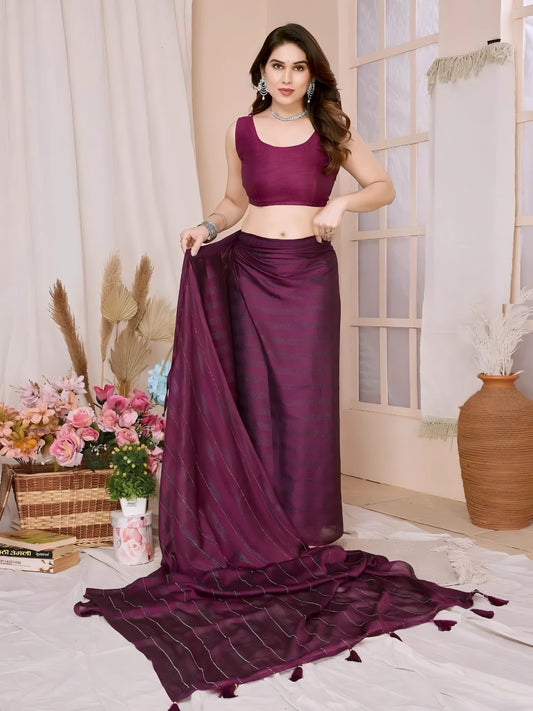 Woman wearing a plum-colored saree with subtle sequin stripes and a sleeveless blouse, accessorized with silver jewelry, standing in a modern interior decorated with vases of pink and blue flowers, a wicker basket, and neutral decor elements