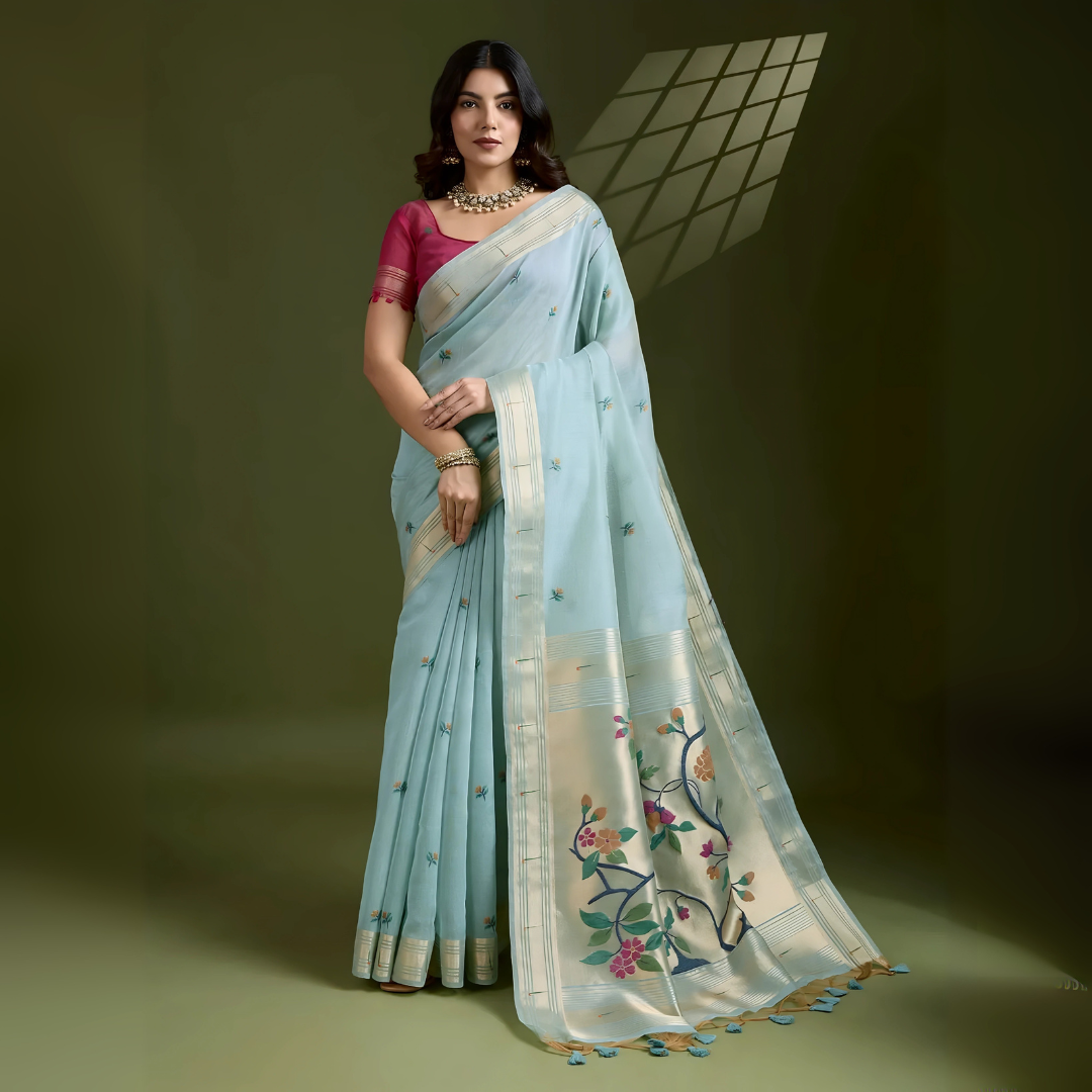 Woman wearing a powder blue silk saree with an off-white floral embroidered pallu and gold zari border, paired with a contrasting magenta blouse and traditional jewelry, elegantly posing in a studio with olive green walls and patterned window light.