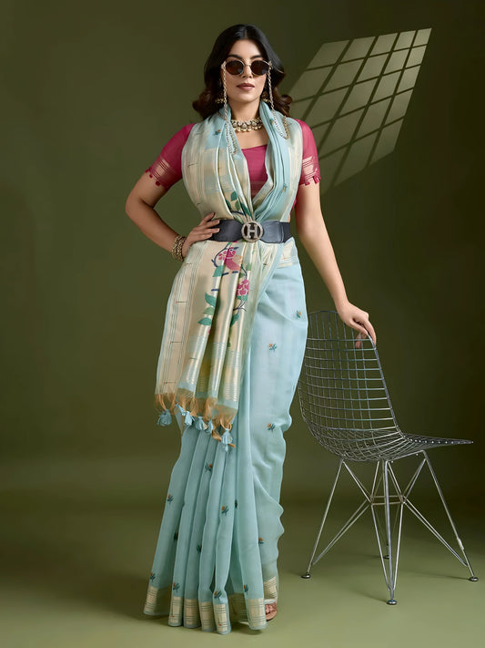 Woman wearing a powder blue silk saree with an off-white floral embroidered pallu and gold zari border, paired with a contrasting magenta blouse and traditional jewelry, elegantly posing in a studio with olive green walls and patterned window light.