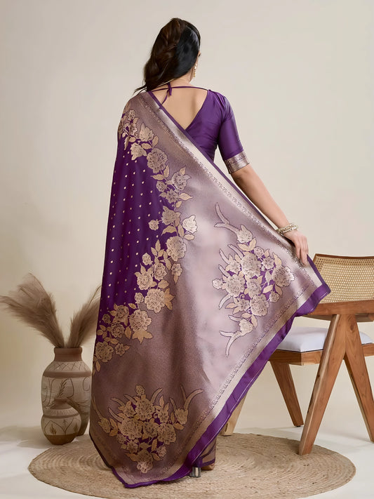 Woman wearing a rich purple Banarasi silk saree with intricate gold floral zari border and pallu, paired with a matching blouse and traditional jewelry, elegantly posing beside a wooden chair in a minimalist studio with neutral decor and ceramic vases.