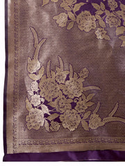 Woman wearing a rich purple Banarasi silk saree with intricate gold floral zari border and pallu, paired with a matching blouse and traditional jewelry, elegantly posing beside a wooden chair in a minimalist studio with neutral decor and ceramic vases.