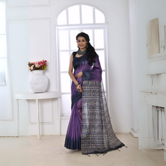 Woman wearing a purple and black Ikat printed saree with geometric patterns on the pallu, styled with a matching blouse and statement necklace, standing in a modern white interior with large arched windows and a white console table with a floral vase.