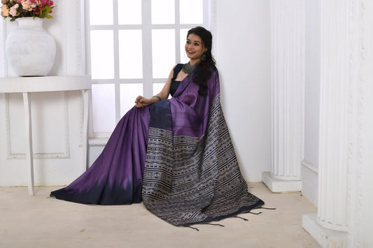 Woman wearing a purple and black Ikat printed saree with geometric patterns on the pallu, styled with a matching blouse and statement necklace, standing in a modern white interior with large arched windows and a white console table with a floral vase.