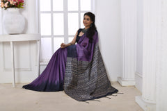 Woman wearing a purple and black Ikat printed saree with geometric patterns on the pallu, styled with a matching blouse and statement necklace, standing in a modern white interior with large arched windows and a white console table with a floral vase.