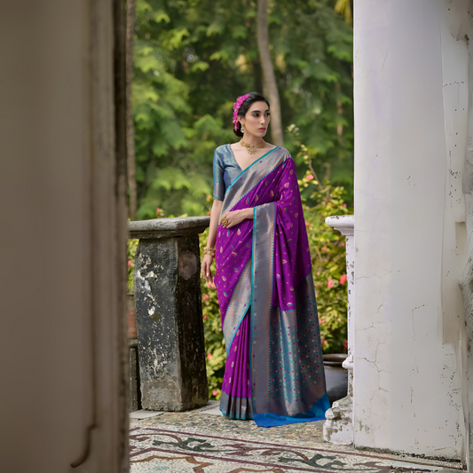 Woman wearing a purple and blue silk saree with silver zari border and polka dot motifs, paired with a blue blouse, standing on a vintage veranda with lush green trees in the background.