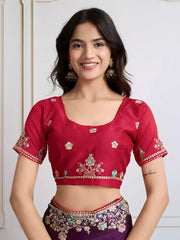 Woman wearing a deep purple chiffon saree featuring delicate floral embroidery and an ornate embroidered border, paired with a short-sleeve magenta blouse. She stands elegantly in a classic white paneled room with a vintage rotary telephone on a carved wooden side table, a modern wooden armchair with a woven seat, and a richly patterned rug for a sophisticated, traditional look.