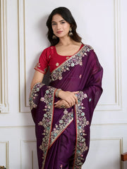 Woman wearing a deep purple chiffon saree featuring delicate floral embroidery and an ornate embroidered border, paired with a short-sleeve magenta blouse. She stands elegantly in a classic white paneled room with a vintage rotary telephone on a carved wooden side table, a modern wooden armchair with a woven seat, and a richly patterned rug for a sophisticated, traditional look.