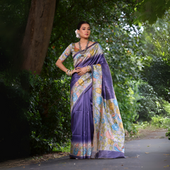 Woman wearing a purple saree with vibrant Madhubani-inspired floral print border and pallu, paired with a matching blouse and traditional jewelry, standing gracefully on a garden pathway surrounded by lush green trees and foliage.