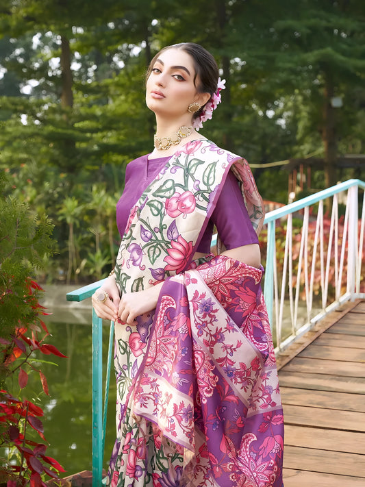 Woman wearing a purple saree with vibrant Madhubani-inspired floral print border and pallu, paired with a matching blouse and traditional jewelry, gracefully standing on stone steps beside a wooden bridge surrounded by lush greenery and colorful garden plants.