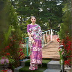 Woman wearing a purple saree with vibrant Madhubani-inspired floral print border and pallu, paired with a matching blouse and traditional jewelry, gracefully standing on stone steps beside a wooden bridge surrounded by lush greenery and colorful garden plants.