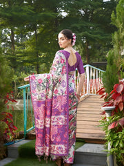 Woman wearing a purple saree with vibrant Madhubani-inspired floral print border and pallu, paired with a matching blouse and traditional jewelry, gracefully standing on stone steps beside a wooden bridge surrounded by lush greenery and colorful garden plants.