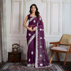 Woman wearing a deep purple organza saree featuring intricate white floral embroidery and a matching embroidered border, paired with a sleeveless blouse. She is elegantly posed in a classic white paneled room with a vintage rotary telephone on a carved wooden side table and a modern wooden chair with a woven seat, all atop an ornate patterned rug for a sophisticated traditional ambiance.