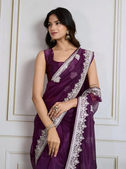Woman wearing a deep purple organza saree featuring intricate white floral embroidery and a matching embroidered border, paired with a sleeveless blouse. She is elegantly posed in a classic white paneled room with a vintage rotary telephone on a carved wooden side table and a modern wooden chair with a woven seat, all atop an ornate patterned rug for a sophisticated traditional ambiance.