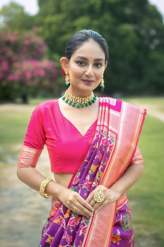  Woman wearing a vibrant purple silk saree with pink borders and multicolor floral motifs, paired with a pink blouse and traditional jewelry, standing gracefully on a garden path surrounded by lush greenery and blooming trees.