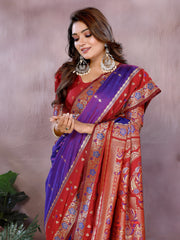 Woman wearing a purple and red Banarasi saree with an ornate golden border and intricate woven motifs, paired with a matching red blouse adorned with gold detailing. She poses gracefully in a modern studio setting featuring blush-toned vases, potted green plants, and a neutral pink backdrop for an elegant traditional look.
