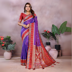 Woman wearing a purple and red Banarasi saree with an ornate golden border and intricate woven motifs, paired with a matching red blouse adorned with gold detailing. She poses gracefully in a modern studio setting featuring blush-toned vases, potted green plants, and a neutral pink backdrop for an elegant traditional look.