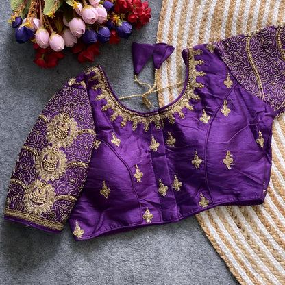  Traditional purple silk blouse featuring intricate gold embroidery and beadwork with floral and paisley motifs on the sleeves and neckline, displayed flat on a textured surface with a bunch of artificial flowers and a woven mat in the background.
