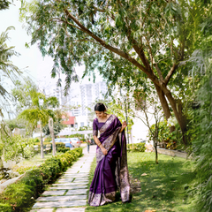 Woman wearing a rich purple silk saree with intricate gold border and matching blouse, standing gracefully on a stone pathway in a lush green garden with manicured lawns, trees, and city buildings in the background.