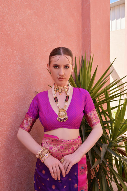 Woman wearing a purple silk saree with pink and gold border, adorned with traditional gold jewelry, standing by a terracotta wall and green plantv.