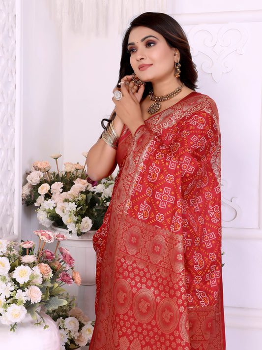 Woman draped in a vibrant red Banarasi bandhani saree featuring intricate white tie-dye patterns and an ornate zari border, paired with a matching sleeveless blouse and silver jewelry. She poses elegantly in a classic white room decorated with decorative wall panels and lush flower arrangements in white vases.