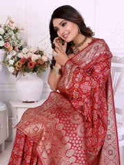Woman draped in a vibrant red Banarasi bandhani saree featuring intricate white tie-dye patterns and an ornate zari border, paired with a matching short-sleeve blouse and traditional jewelry. She stands gracefully in a classic white room adorned with paneled walls and elegant white vases filled with pastel and rose-toned artificial flower arrangements.