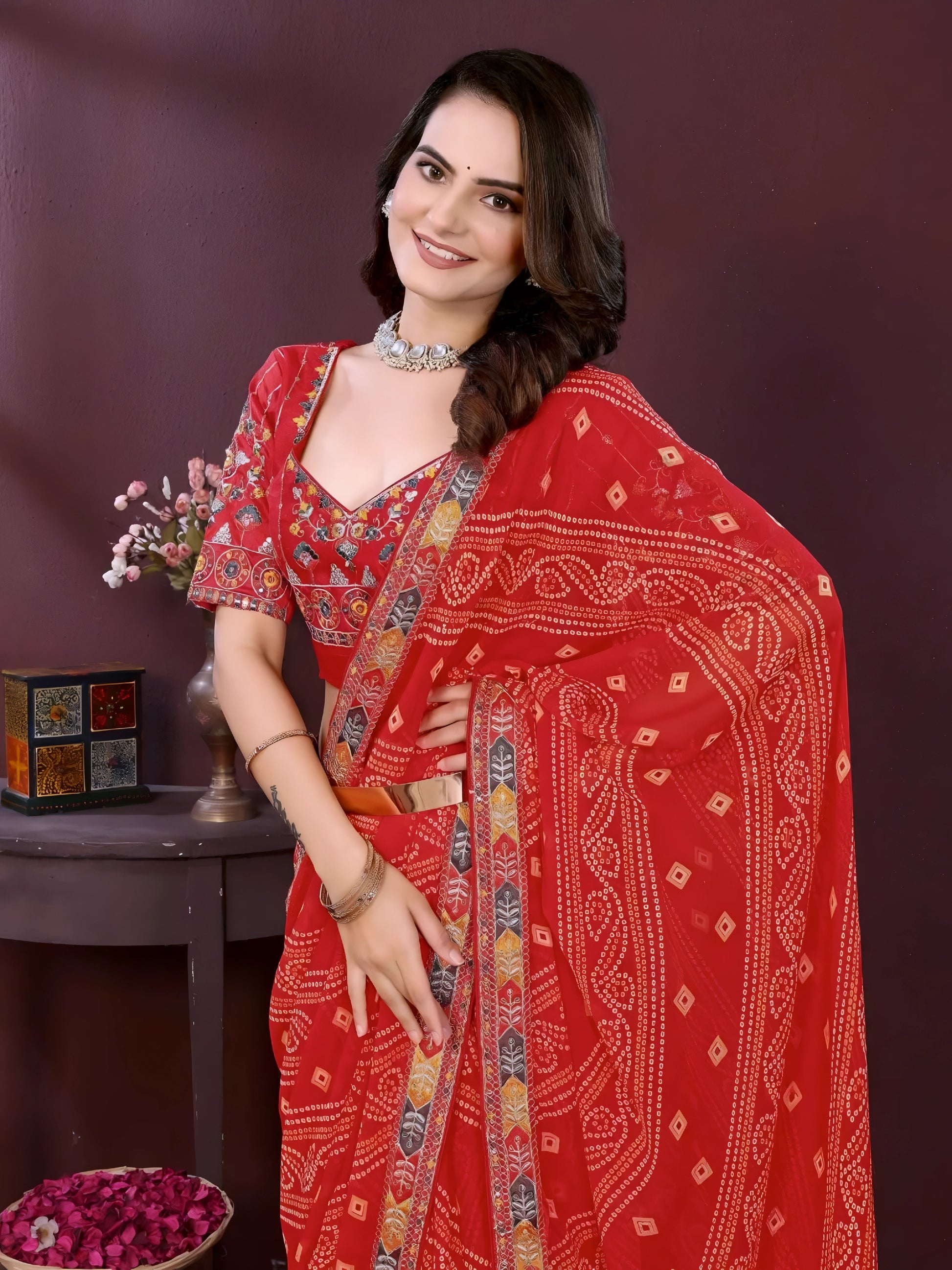 Woman wearing a red bandhani print saree with a contrast patterned border and matching blouse, styled with a choker necklace in an indoor studio setup, Indian ethnic wear product photo