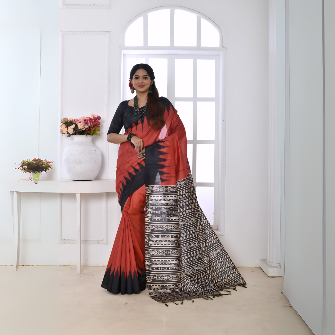  Woman wearing a red and black Ikat printed saree with geometric border and pallu, paired with a black blouse, standing in a modern white interior with a window and floral decor on a console table.