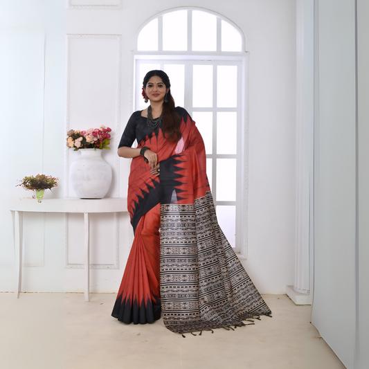  Woman wearing a red and black Ikat printed saree with geometric border and pallu, paired with a black blouse, standing in a modern white interior with a window and floral decor on a console table.