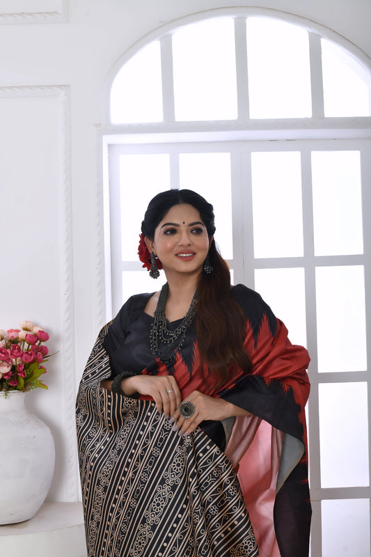 Woman wearing a red and black Ikat printed saree with geometric border and pallu, paired with a black blouse, standing in a modern white interior with a window and floral decor on a console table.